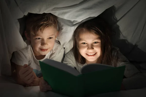 Cute little brother and sister reading bedtime story under blanket in evening — Stock Photo, Image