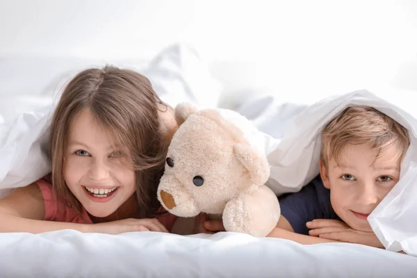 Porträtt av glada lilla bror och syster liggande på sängen under täcket — Stockfoto