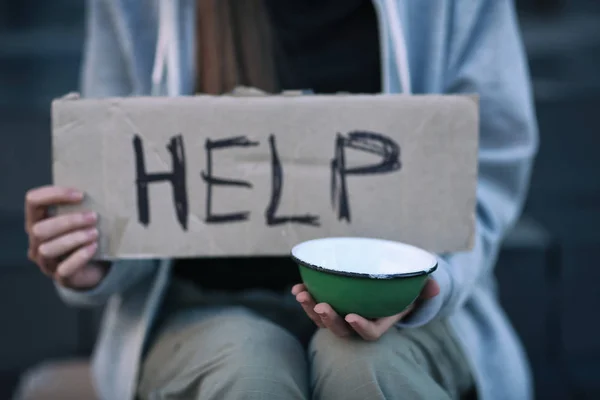 Homeless poor woman holding empty bowl and piece of cardboard with word HELP outdoors, closeup — Stock Photo, Image