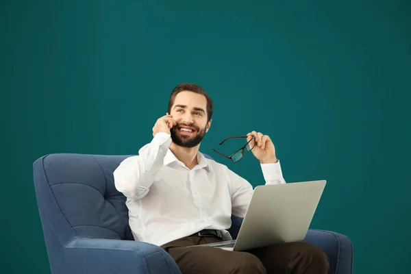 Handsome man with laptop talking on mobile phone while sitting in comfortable armchair against color background — Stock Photo, Image