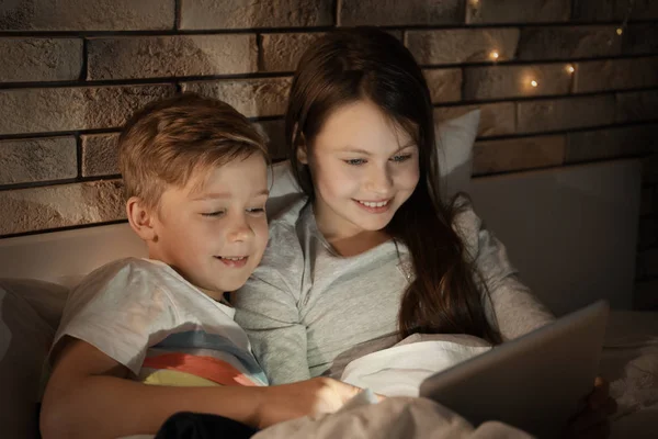Симпатичные младшие брат и сестра читают сказку на ночь с планшетного компьютера вечером — стоковое фото
