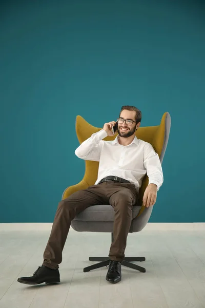 Jóképű férfi beszél, mobiltelefon színes falnak kényelmes karosszékben ülve — Stock Fotó