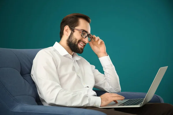 Schöner Mann mit Laptop sitzt im bequemen Sessel vor farbigem Hintergrund — Stockfoto