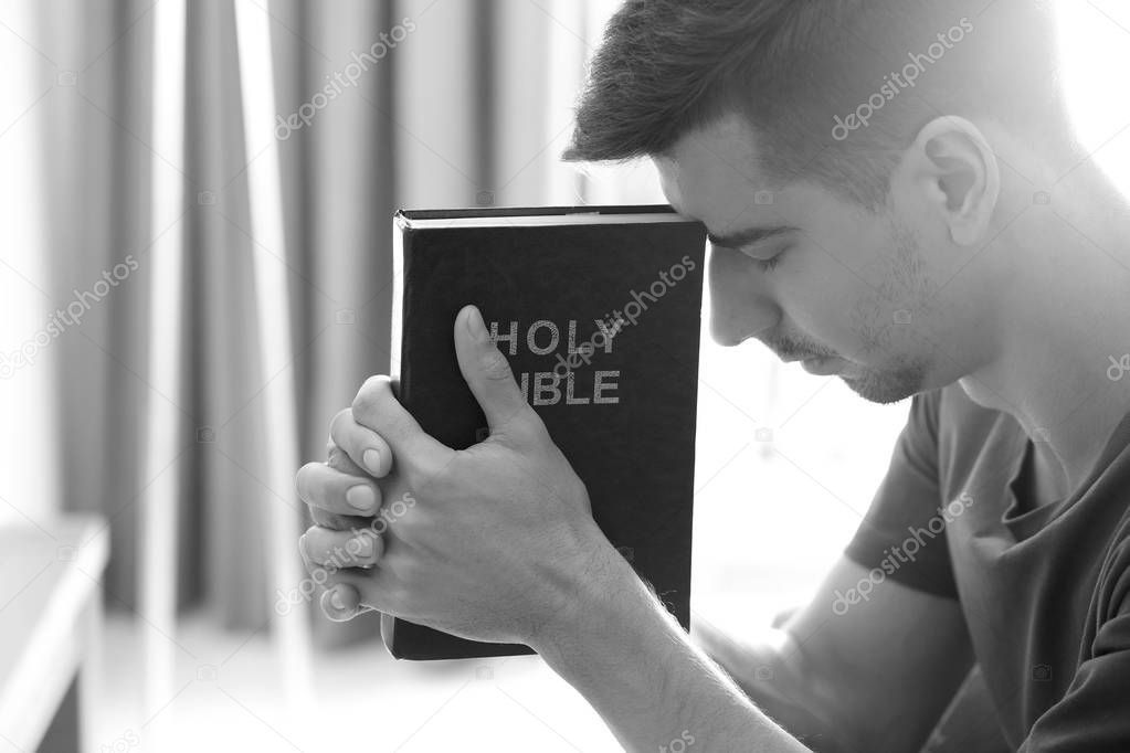 Religious young man with Bible praying at home, toned in black and white
