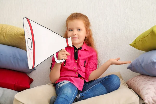 Nettes kleines Mädchen mit einem Megafon aus Papier sitzt zwischen einem Stapel Kissen im Haus — Stockfoto