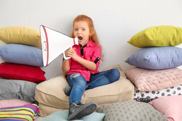 Симпатична маленька дівчинка з паперовим мегафоном сидить на купі подушок у приміщенні — стокове фото