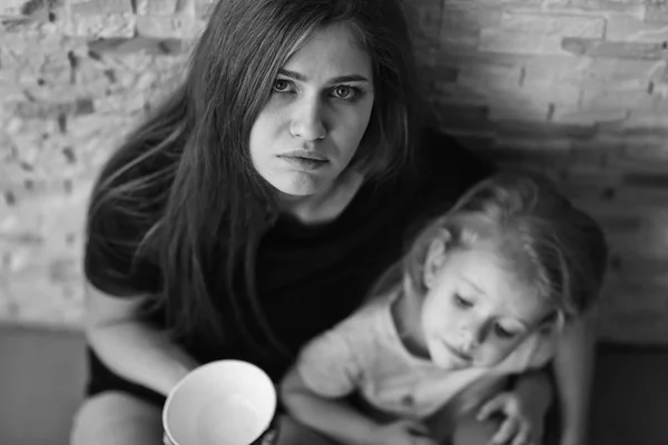 Homeless poor woman with little daughter holding empty bowl and asking alms near brick wall — Stock Photo, Image