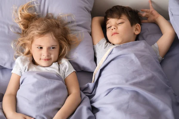 Lindo chico durmiendo mientras su hermana pequeña tratando de conciliar el sueño en la cama en casa. Hora de dormir familiar — Foto de Stock