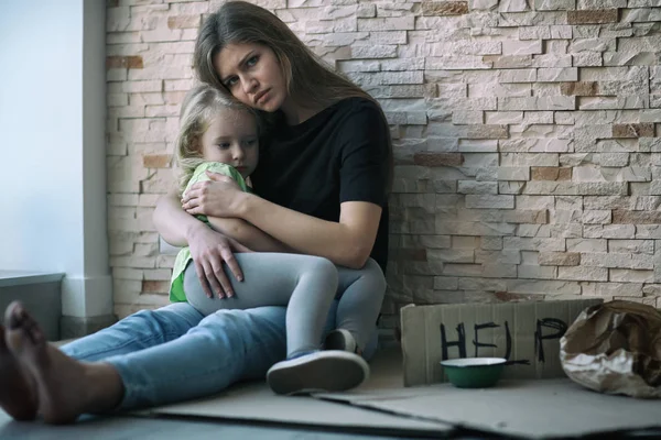 Sin hogar pobre mujer y su pequeña hija sentada cerca de la pared de ladrillo y pidiendo ayuda — Foto de Stock