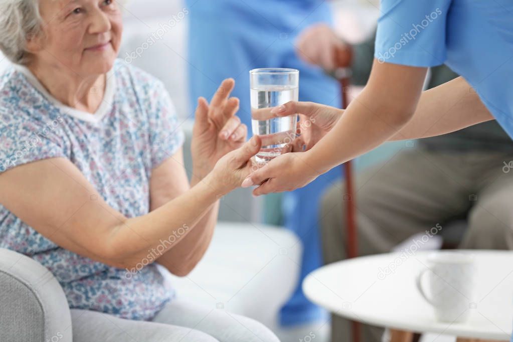 Young caregiver giving glass of water to senior woman indoors