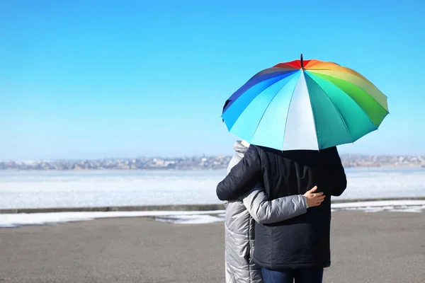 Jovem casal romântico com guarda-chuva colorido na margem do rio — Fotografia de Stock