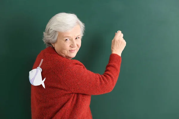 Professora sénior com peixe de papel nas costas a escrever em quadro-negro. Dia do tolo de abril celebração — Fotografia de Stock