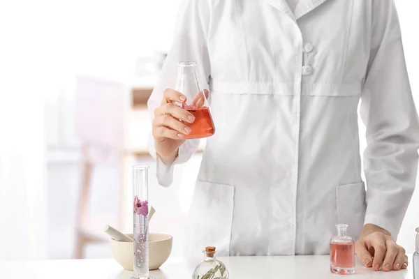 Woman in lab coat holding glass flask with perfume oil near table indoors — Stock Photo, Image