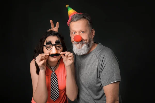 Young woman and mature man in funny disguise on dark background. April fool's day celebration — Stock Photo, Image