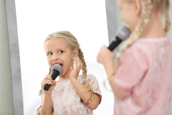 Nettes kleines Mädchen mit Mikrofon posiert vor Spiegel drinnen — Stockfoto