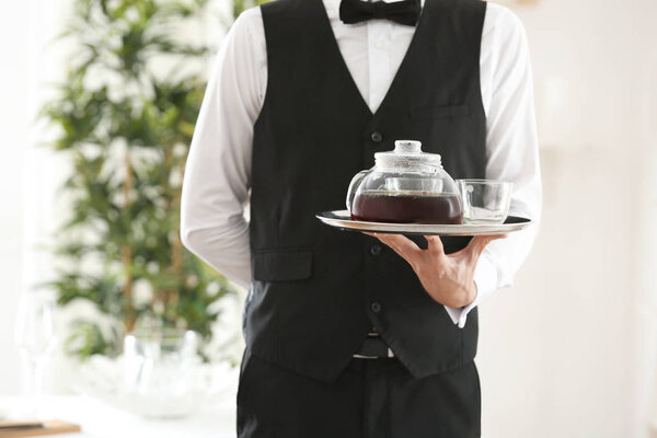 Официант держит поднос со стеклянным чайником и чашками в ресторане
