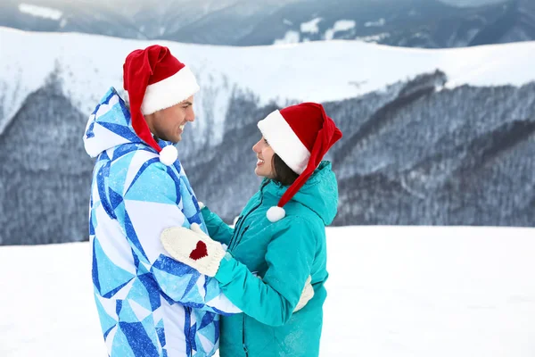 Lyckliga paret i varma vantar och Santa hattar på snöiga resort. Vintersemester — Stockfoto