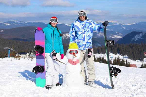 Pareja feliz con muñeco de nieve y tablas de snowboard en la estación de esquí. Vacaciones de invierno —  Fotos de Stock