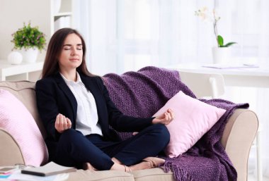 Evdeki kanepede meditasyon yapan genç bir kadın.