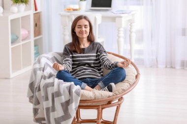 Young woman meditating in lounge chair at home clipart