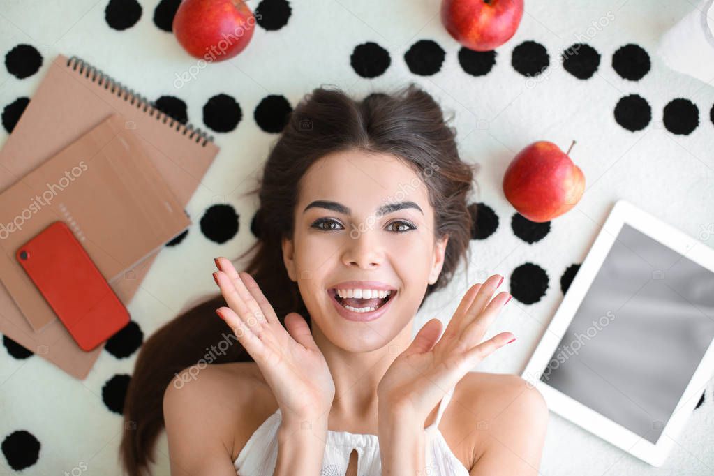 Beautiful young woman with tablet and apples lying on carpet, top view