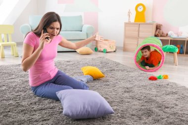 Playful little boy and annoyed nanny talking on phone at home clipart