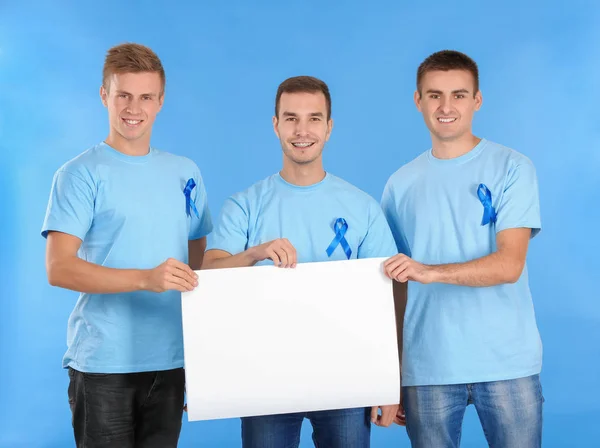Young men in t-shirts with blue ribbons holding blank banner on color background. Prostate cancer awareness concept