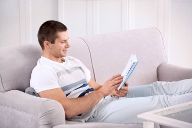 Genç adam evdeki kanepede kitap okuyor.
