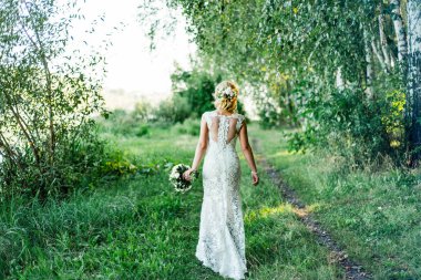 Gelin bir buket ile beyaz elbiseli ormandaki bir yolda yürüyor