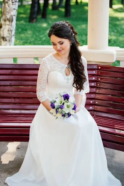 Hermosa novia en vestido blanco con ramo en las manos se sienta en un banco de madera — Foto de Stock