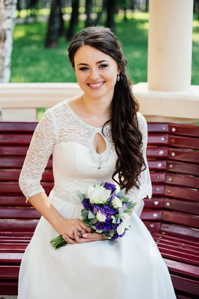 Hermosa novia en vestido blanco con ramo en las manos se sienta en un banco de madera — Foto de Stock