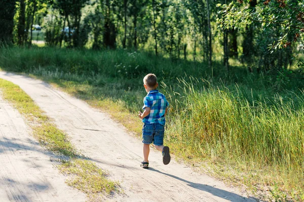 Μικρό αγόρι σε ένα μπλε καρό πουκάμισο και σορτς που τρέχει σε ένα μονοπάτι στο ξύλο — Φωτογραφία Αρχείου