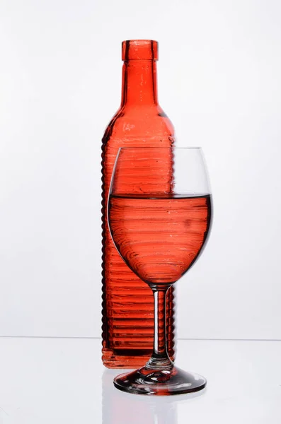 Copa de vino transparente con agua frente a una botella roja sobre un fondo blanco — Foto de Stock