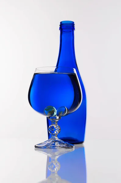 Copa de vino transparente con agua frente a una botella azul sobre un fondo blanco — Foto de Stock