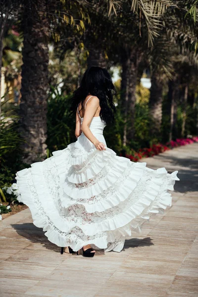 Beautiful girl with long curly hair in a long white dress spinning next to palm trees. Blowing up dress — Stock Photo, Image