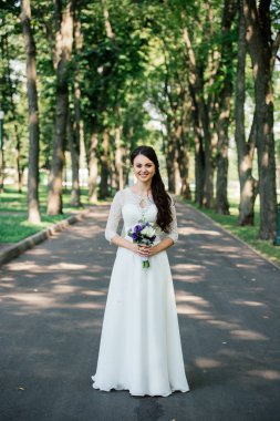 Güzel genç gülümseyen esmer gelin gelinlik yeşil yaprakların arka plan açık havada ellerinde çiçekler buket