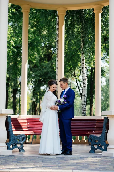 La novia y el novio tomados de la mano cerca del banco de madera en el parque. Pareja de boda enamorada en el día de la boda — Foto de Stock