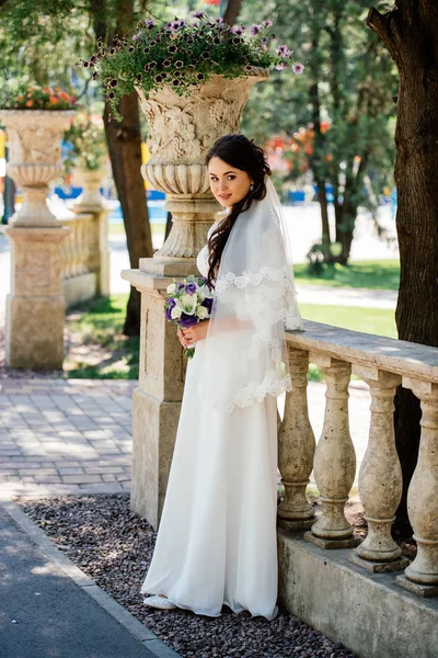 Hermosa joven morena sonriente novia en vestido de novia con ramo de flores en las manos al aire libre en el fondo de hojas verdes — Foto de Stock