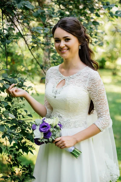 Bella giovane sposa bruna sorridente in abito da sposa con mazzo di fiori in mano all'aperto sullo sfondo di foglie verdi — Foto Stock