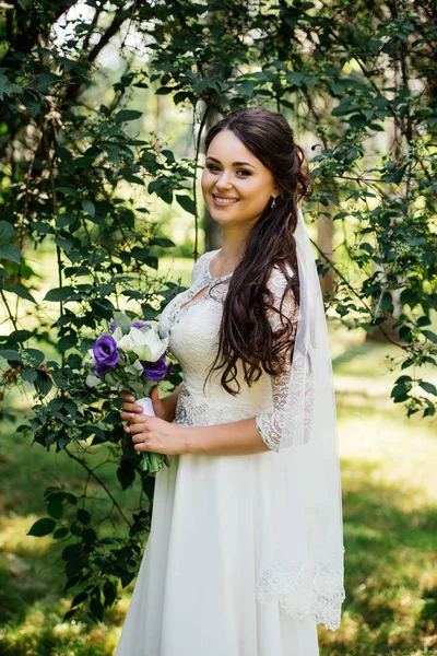Mooie jonge lachende brunette bruid in trouwjurk met boeket bloemen in handen buiten op de achtergrond van groene bladeren — Stockfoto