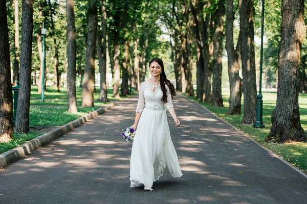 Belle jeune mariée brune souriante en robe de mariée avec bouquet de fleurs dans les mains va dans le parc à l'extérieur — Photo