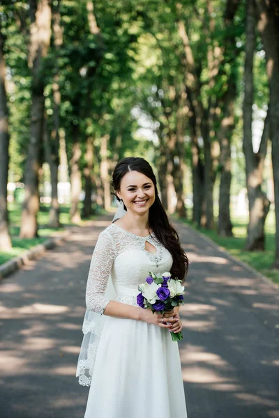 Mooie jonge lachende brunette bruid in trouwjurk met boeket bloemen in handen buiten op de achtergrond van groene bladeren — Stockfoto