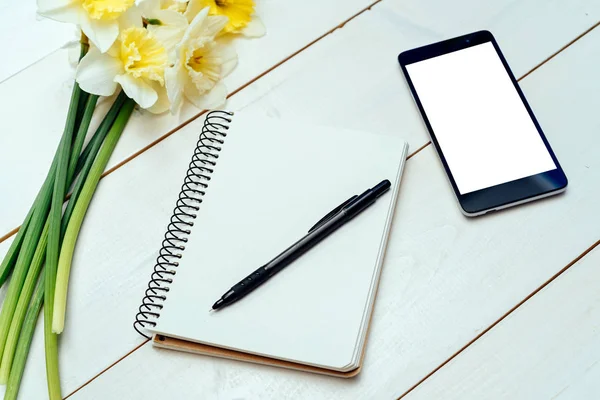 Nergis Beyaz ahşap zemin üzerine sarı buket. Boş kart düz yatıyordu. Nergis, boş günlüğü, kalem ve telefon, boş alan tablo üstten görünüm