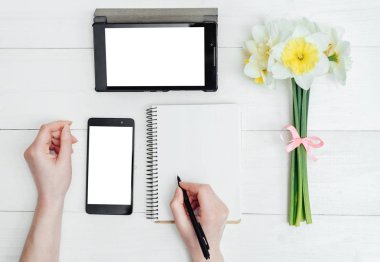 Üstten görünüm nergis, boş günlüğü, tablet, telefon ve kızın elleri kalem, boş alan ile tablo. Kızın ellerini günlüğüne yazma