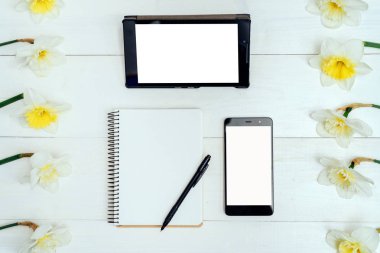 Üstten görünüm nergis, boş günlüğü, kalem, tablet ve telefon, boş alan tablo. Beyaz ahşap zemin üzerine sarı nergis. Boş kart düz yatıyordu