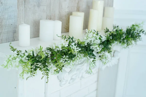 White decorative fireplace with candles on it near wooden wall. Floral decoration of white flowers and greenery over white fireplace