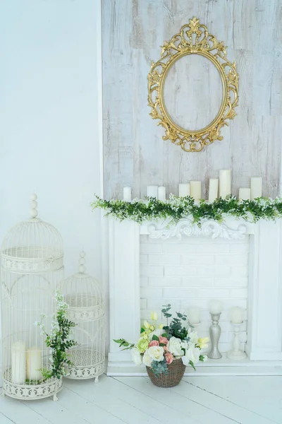 White decorative fireplace with candles and floral decoration on it. Flower arrangement in a basket by the fireplace. Decorative metal cages with candles