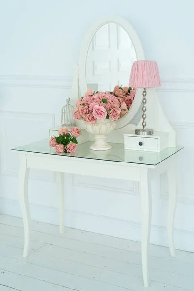 White boudoir table with oval mirror, pink flowers and lamp on it in white room. Detail of the interior of the room with female boudoir