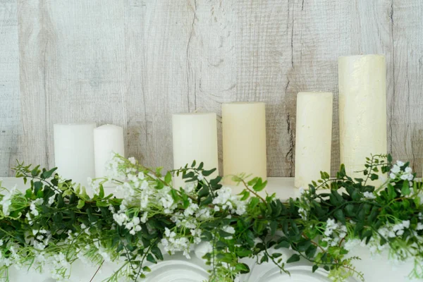 White decorative fireplace with candles on it near wooden wall. Floral decoration of white flowers and greenery over white fireplace, closeup