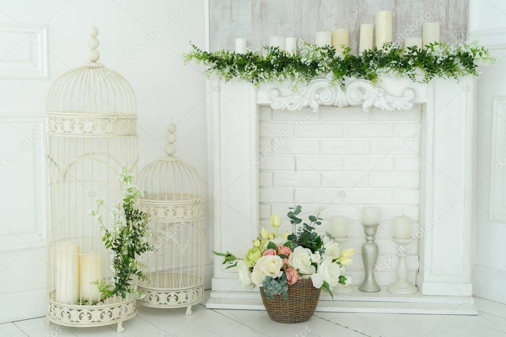 White decorative fireplace with candles and floral decoration on it. Flower arrangement in a basket by the fireplace. Decorative metal cages with candles 